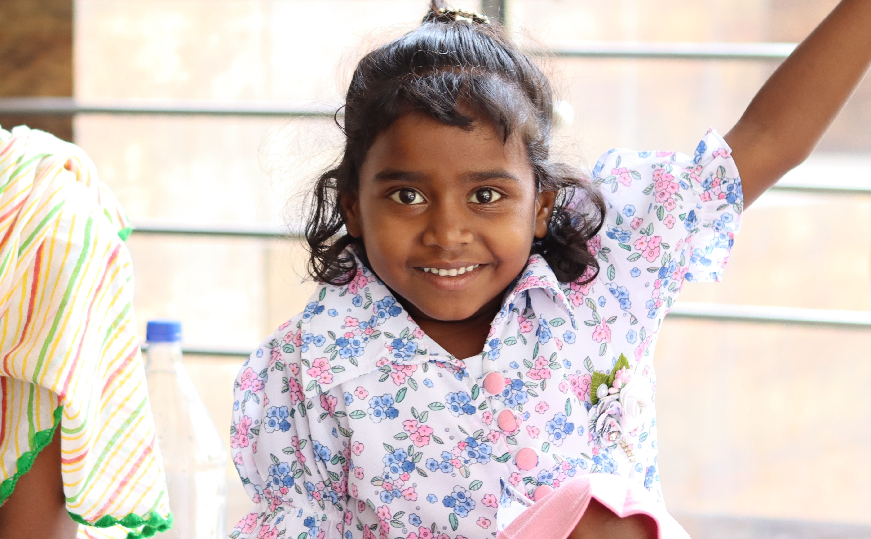 A smiling young girl with dark curly hair, wearing a white dress with pink and blue floral patterns, raises her left arm cheerfully. She is sitting indoors near a railing, and a bottle and colorful clothing are partially visible on the left side.