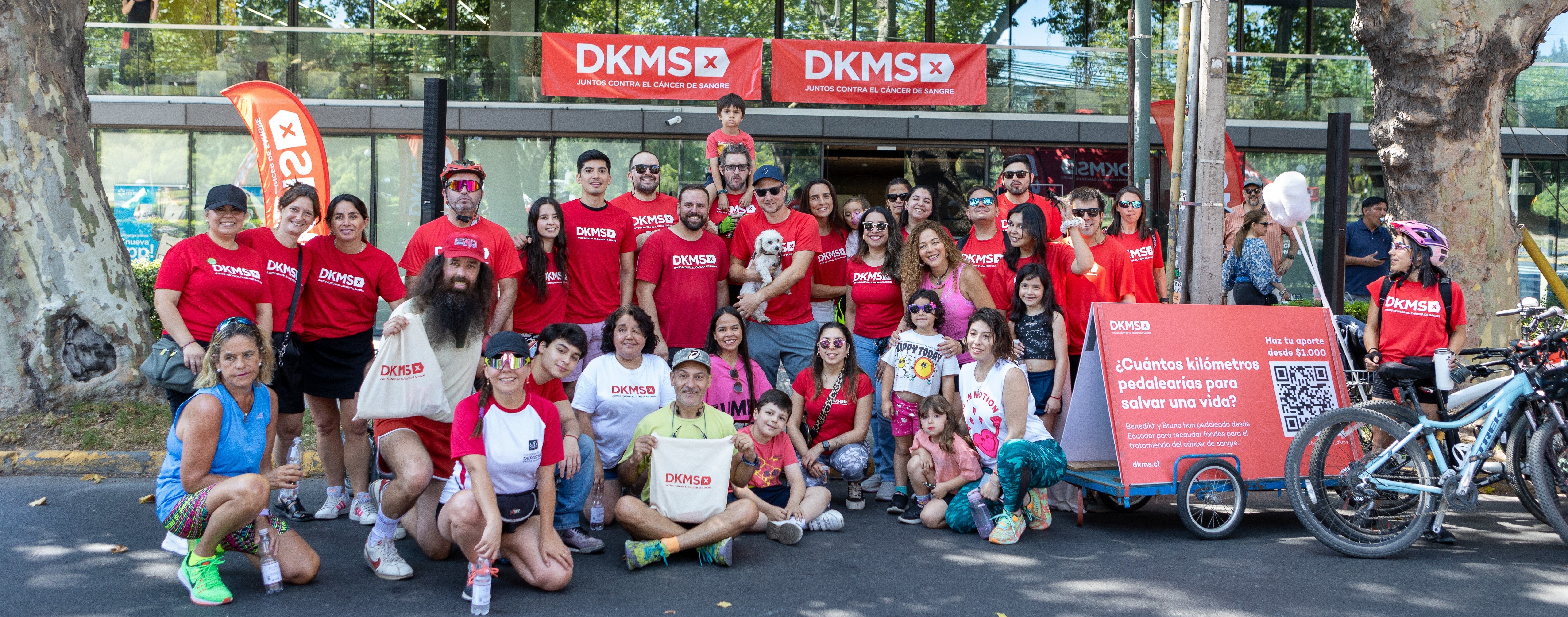 A large, cheerful group of DKMS volunteers, employess, supporters, and families pose together during a community event in Chile. Most are wearing red DKMS shirts and standing in front of a DKMS banner that reads 'Juntos contra el cáncer de sangre', which means 'Together against blood cancer'. A red sign on a bicycle trailer with giant cotton swabs and a QR code asks, '¿Cuántos kilómetros pedalearías para salvar una vida?', which means 'How many kilometers would you cycle to save a life?', encouraging donations and participation in a fundraising bike challenge.