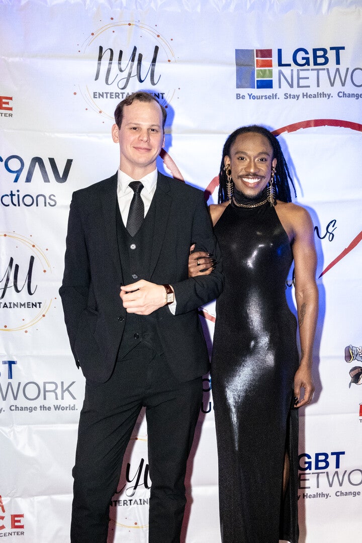 Two people posing together on a red carpet in front of banner. The man on the left is wearing a black suit and tie. The donor Deonté Lee on the right, wrapped his arm around the others man arm, is wearing a shiny black form-fitting, halter-neck gown with statement earrings and a confident smile. The background features logos from LGBT Network, NYU Entertainment, and other sponsors.