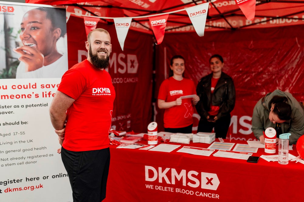 A group of four DKMS volunteers standing at a donor drive booth, all wearing red DKMS shirts. One man stands smiling in front of the booth, while the three others standing under a red DKMS tent behind the table and organizing materials. The booth is decorated with DKMS banners, and a large poster encourages people to register as potential stem cell donors.