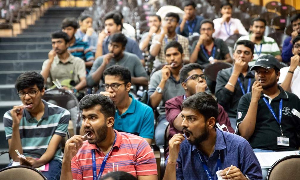 A large group of around 20 young Indian men, sitting in rows in an auditorium, are swabbing the inside of their cheeks with cotton swabs as part of a stem cell donor registration drive. Most are wearing lanyards and casual clothing, and they appear focused on the process.