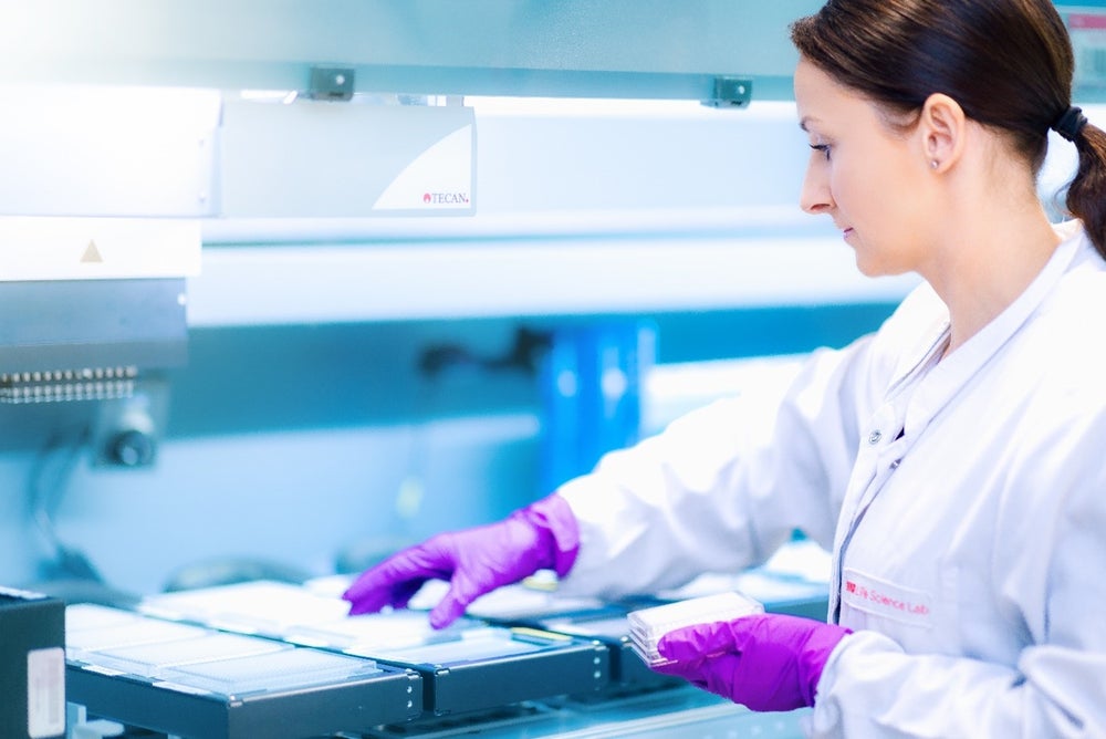 An female employee of the DKMS Life Science Lab with dark brown hair tied back in a ponytail, wearing a white lab coat and purple gloves, works with laboratory equipment.