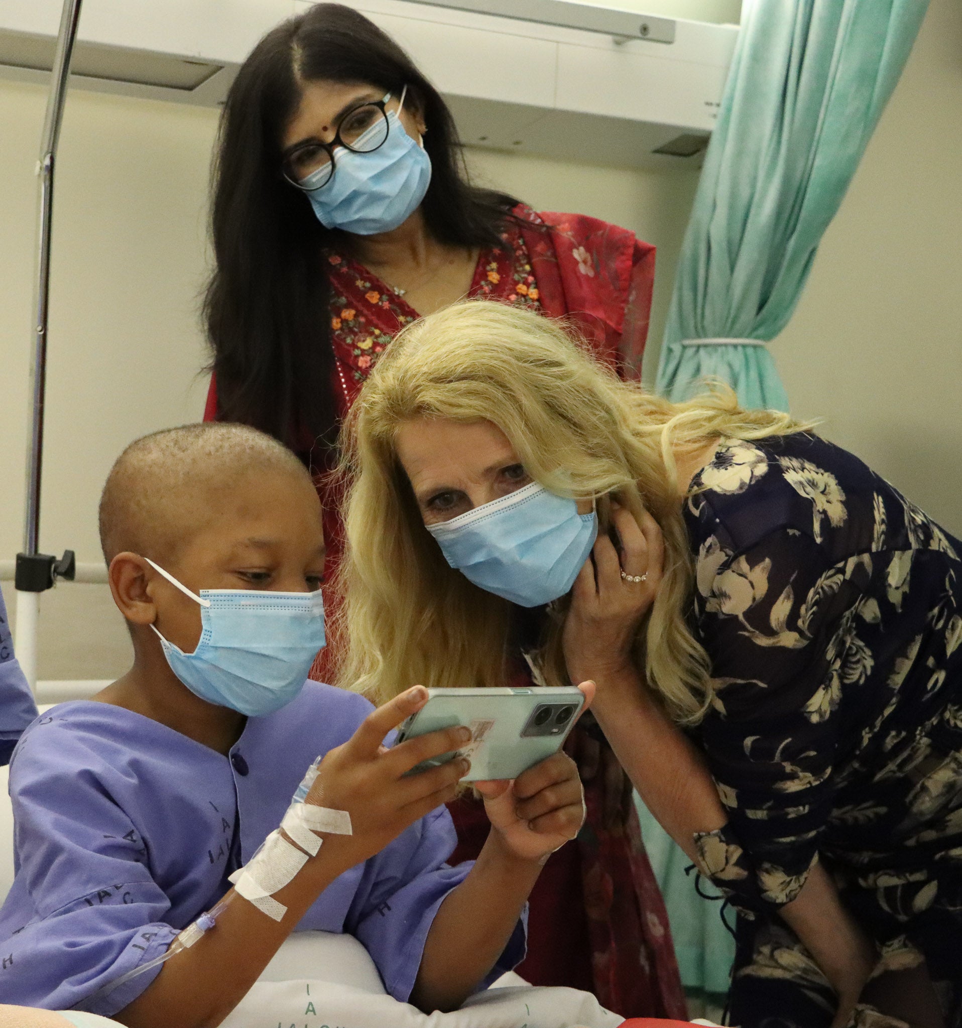 A young male patient, who has a venous access at his arm, wearing a hospital gown and a medical mask, shows something on a smartphone to two women leaning in, also wearing masks. The CEO Elke Neujahr is wearing a black dress with a white floral print and smiling warmly, while the other, wearing glasses and a red outfit, watches from behind. The scene takes place in a hospital room, with a curtain and medical equipment in the background.