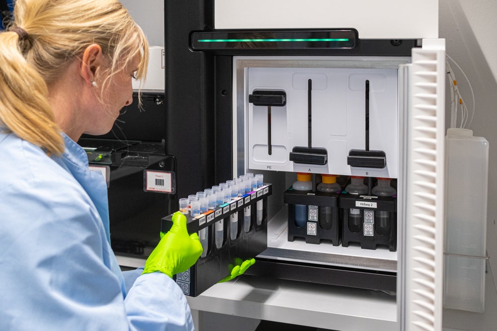 A female lab technician with a blond ponytail, wearing a light blue lab coat and bright green protective gloves, is shown from behind and loading a rack of labeled test tubes into a high-tech laboratory machine. The Lab is equipped with precise instruments and reagents visible inside the machine.