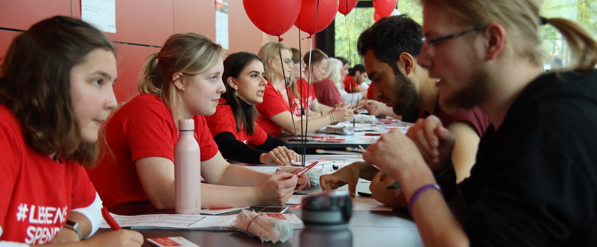 Young DKMS volunteers in red shirts sit at a long table on the left during a university registration event talking to students, which are sitting on the right, about registering as potential donors with DKMS. Swab kits and paperwork are on the table. Red balloons float above the tables, and the atmosphere is focused and energetic as people engage in conversation.