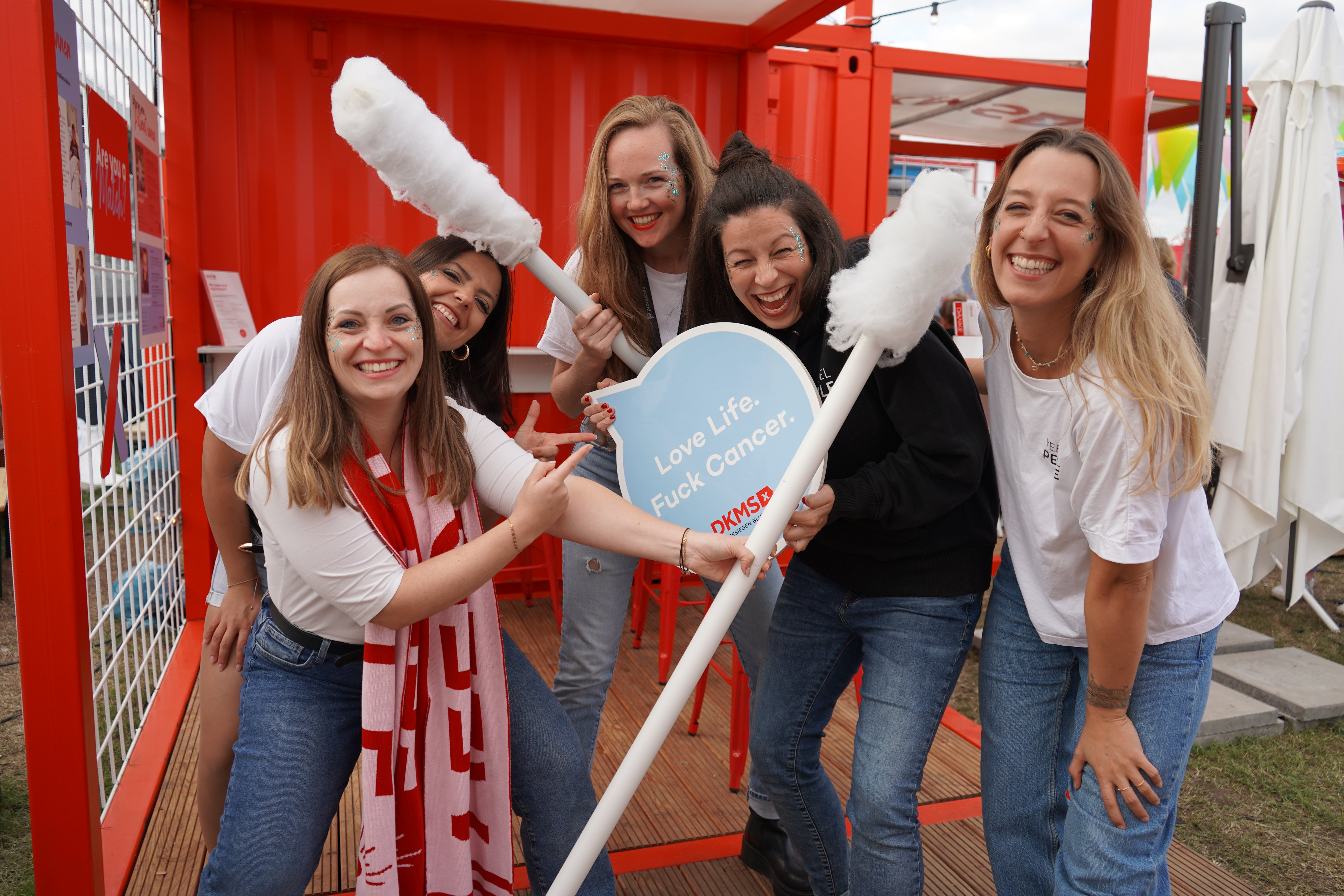 Five smiling women pose in front of a red DKMS booth, holding oversized cotton swabs and a blue speech bubble sign that reads 'Love Life. Fuck Cancer.' They are wearing casual outfits and have glitter on their faces, creating a fun atmosphere.