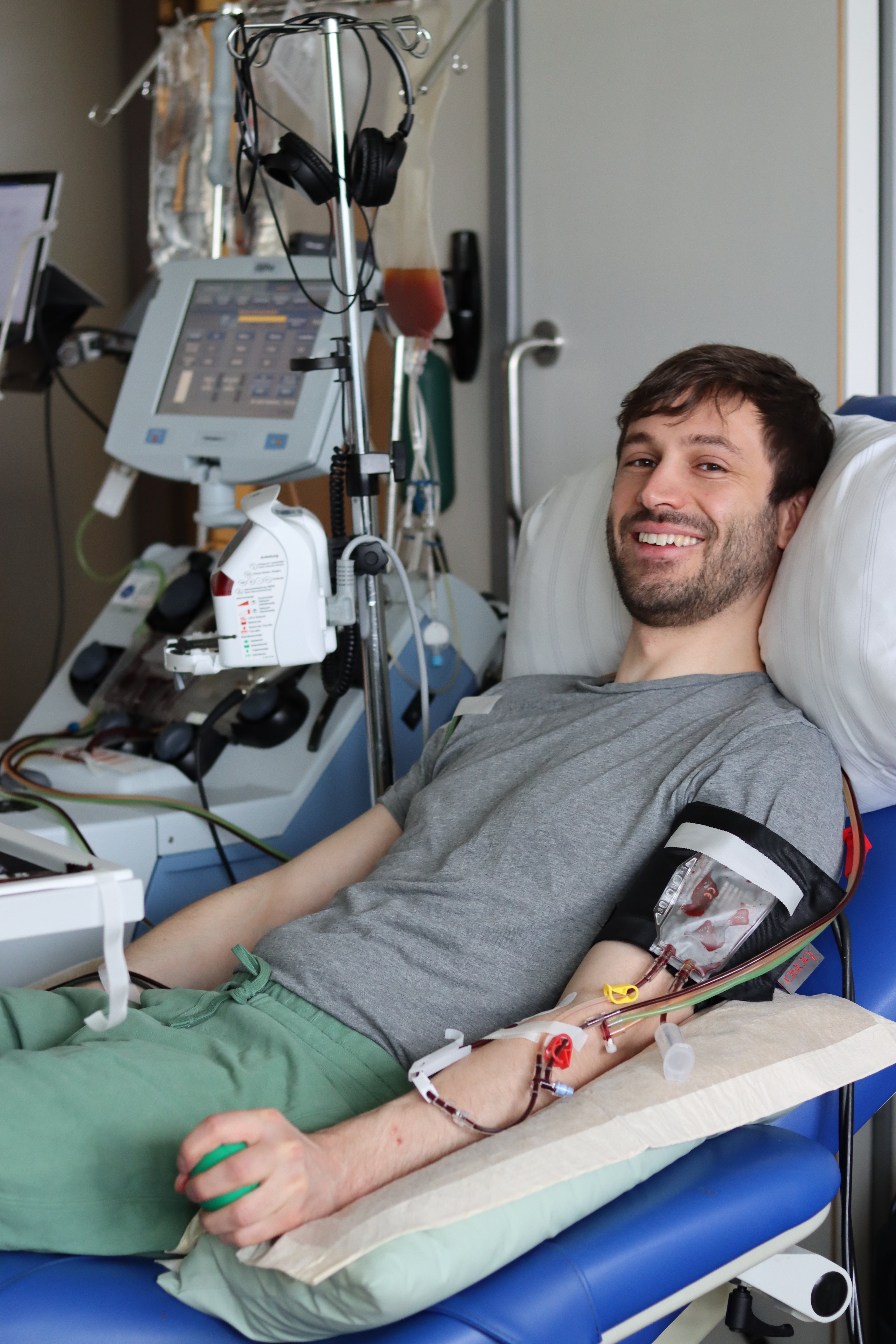 The donor Marvin is smiling and sitting in a chair donating stem cells through apheresis. He is connected to medical equipment, with tubes leading from his left arm to the machine. He is wearing a gray tshirt and green trousers.