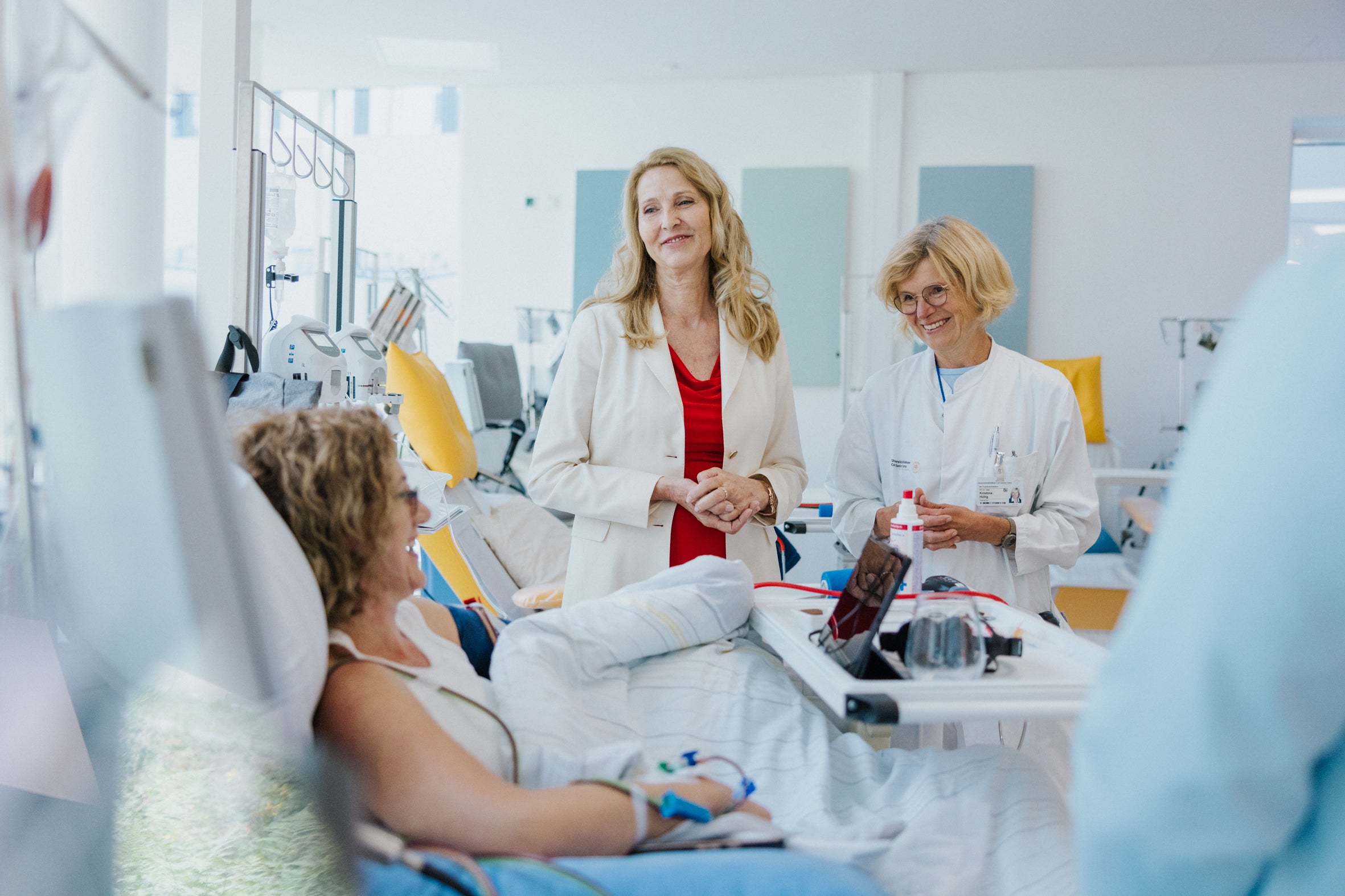 A female stem cell donor lies in a hospital bed during stem cell donation, smiling and talking with two women standing beside her. One woman, wearing a red dress and white blazer, is Dr. Elke Neujahr, Global CEO of DKMS Group. The other, in a white medical coat, is Prof. Dr. Kristina Hölig. The scene takes place in a bright and modern donation facility.