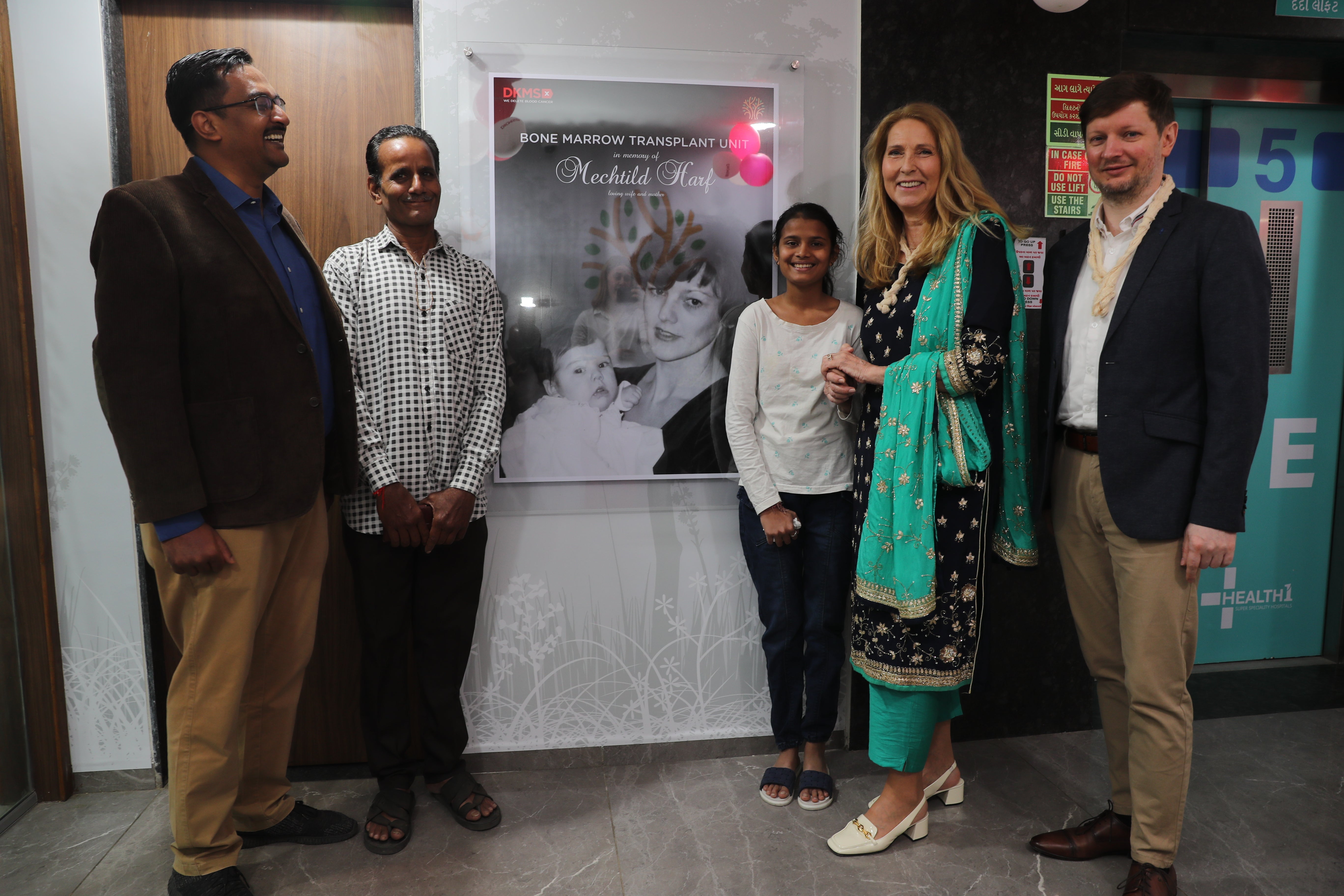 A group of five people, Rajat Kumar Agarwal, President of Sankalp India Foundation, the young girl Tanvi, who was diagnosed with thalassemia, her father, Dr. Elke Neujahr, CEO of DKMS, and Patrick Paul, CEO of DKMS Foundation India, are standing and smiling in front of a poster of Mechtild Harf. They have gathered for the inauguration of the Bone Marrow Transplant Unit in Ahmedabad, India.