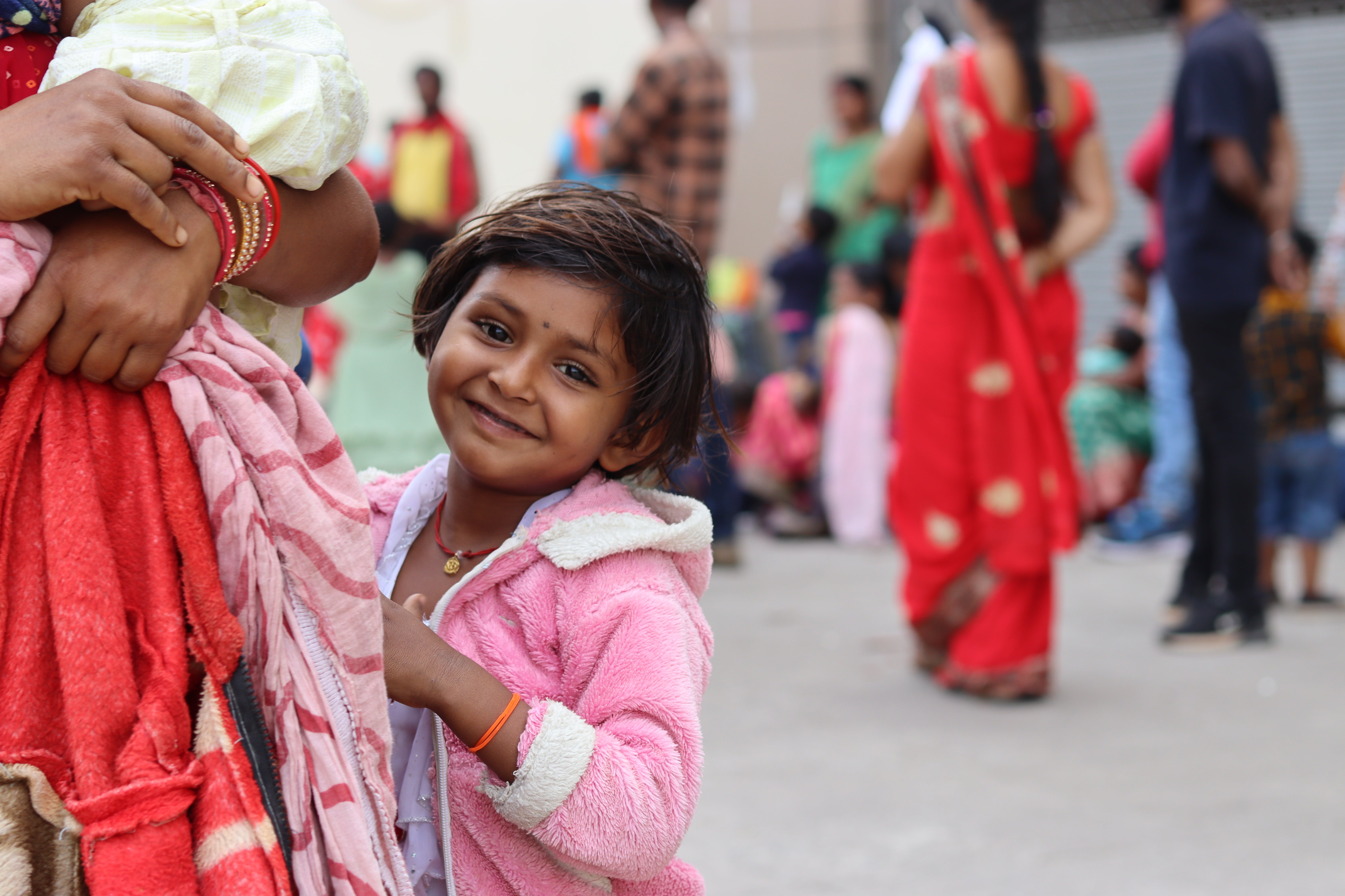 A little Indian girl, who is smiling and wearing a pink hoodie, is standing next to a woman of whom only one arm and the hand are visible. The woman is holding some clothes in her hand. In the background, people in colorful clothing can be seen.