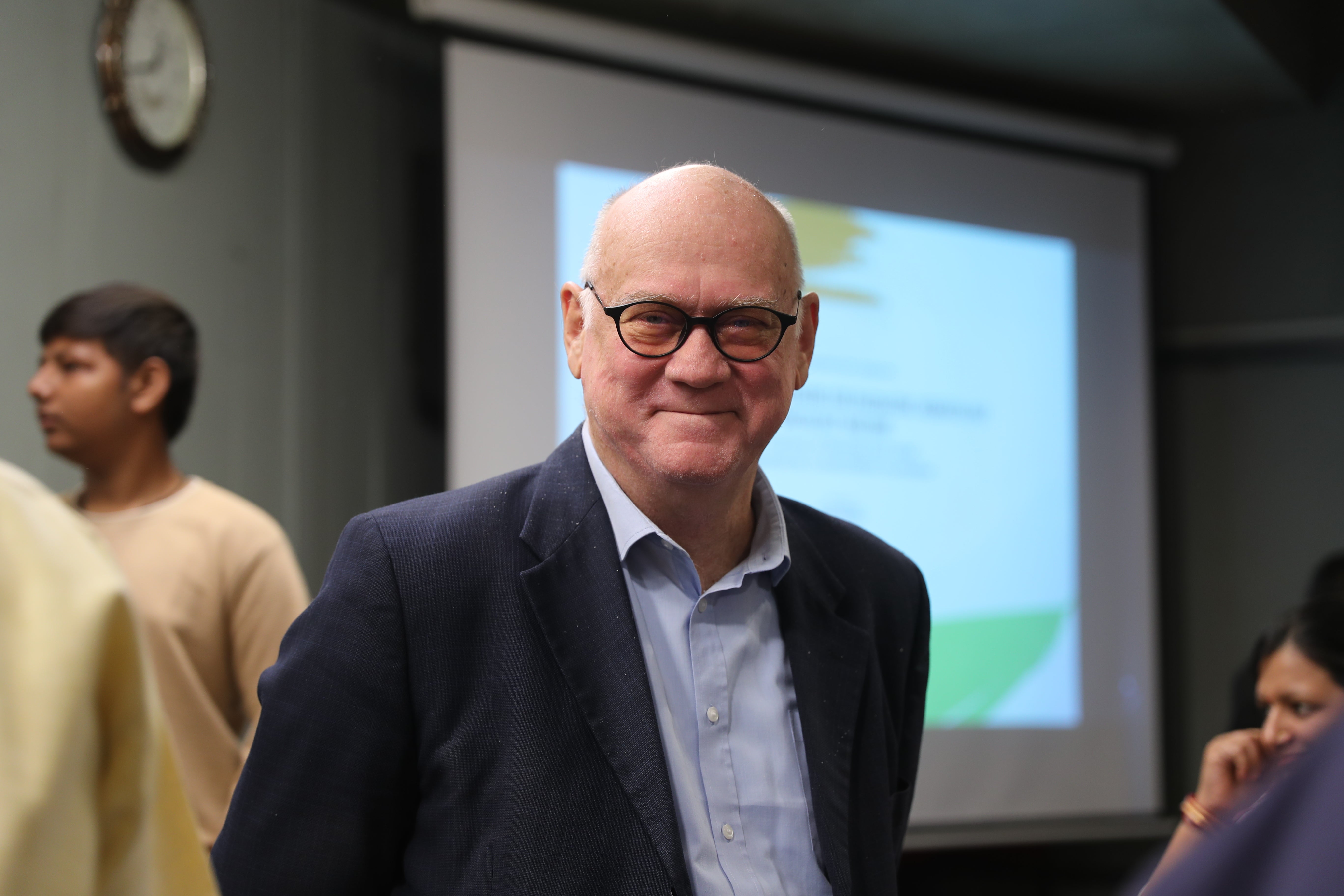 The Cure2Children’s co-founder and pediatric hematologist-oncologist Dr. Lawrence Faulkner is smiling gently at the camera. He is wearing a dark suit and glasses, standing indoors in front of a projection screen. Other people and a presentation are visible in the background.