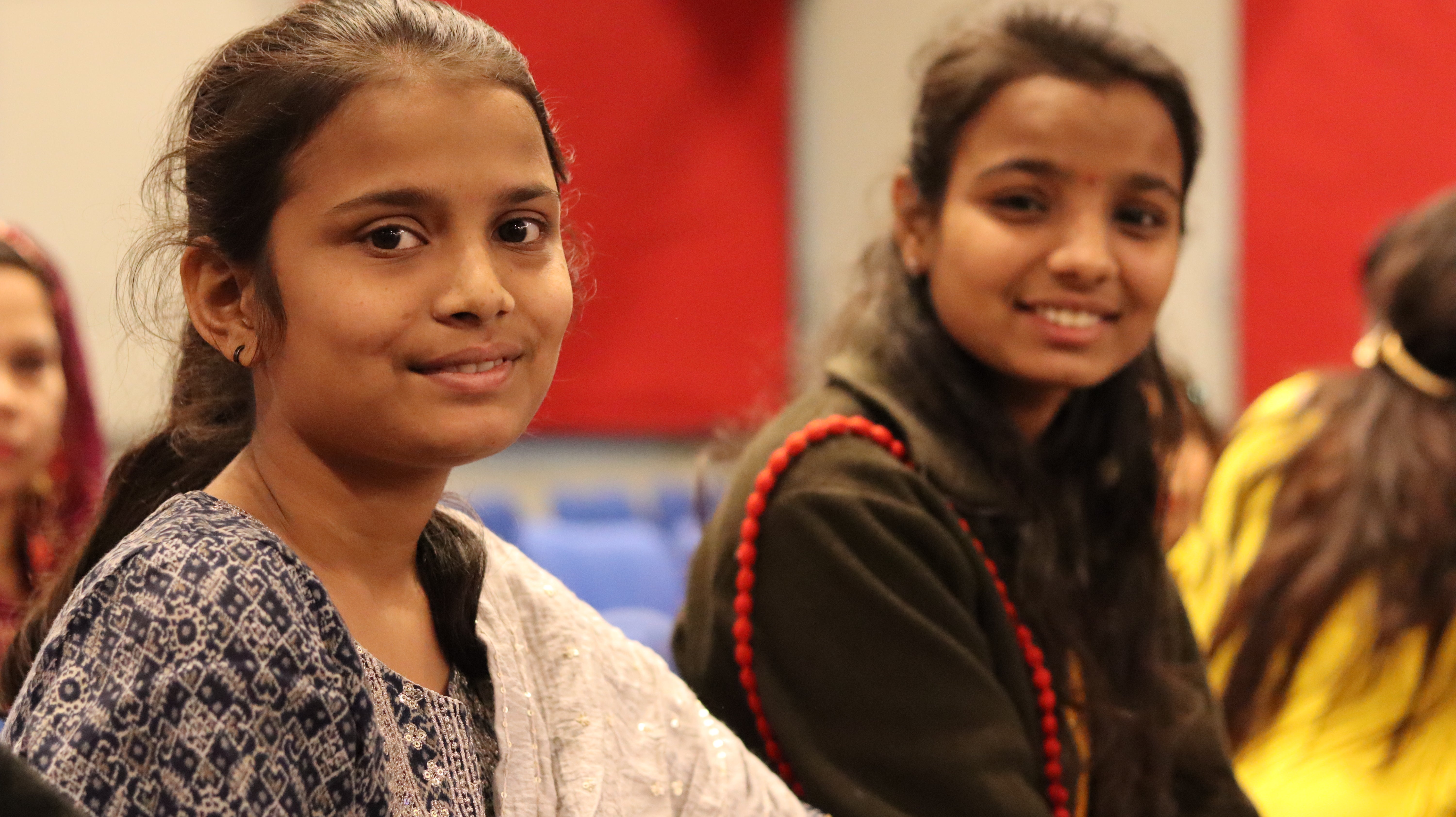 The young girl Tanvi is sitting indoors with her sister. They both have dark tied back hair and are smiling gently at the camera. Tanvi, in the foreground, wears a patterned blue and white shirt, while her sister next to her wears a dark jacket with red accents. Other people are slightly blurred in the background.