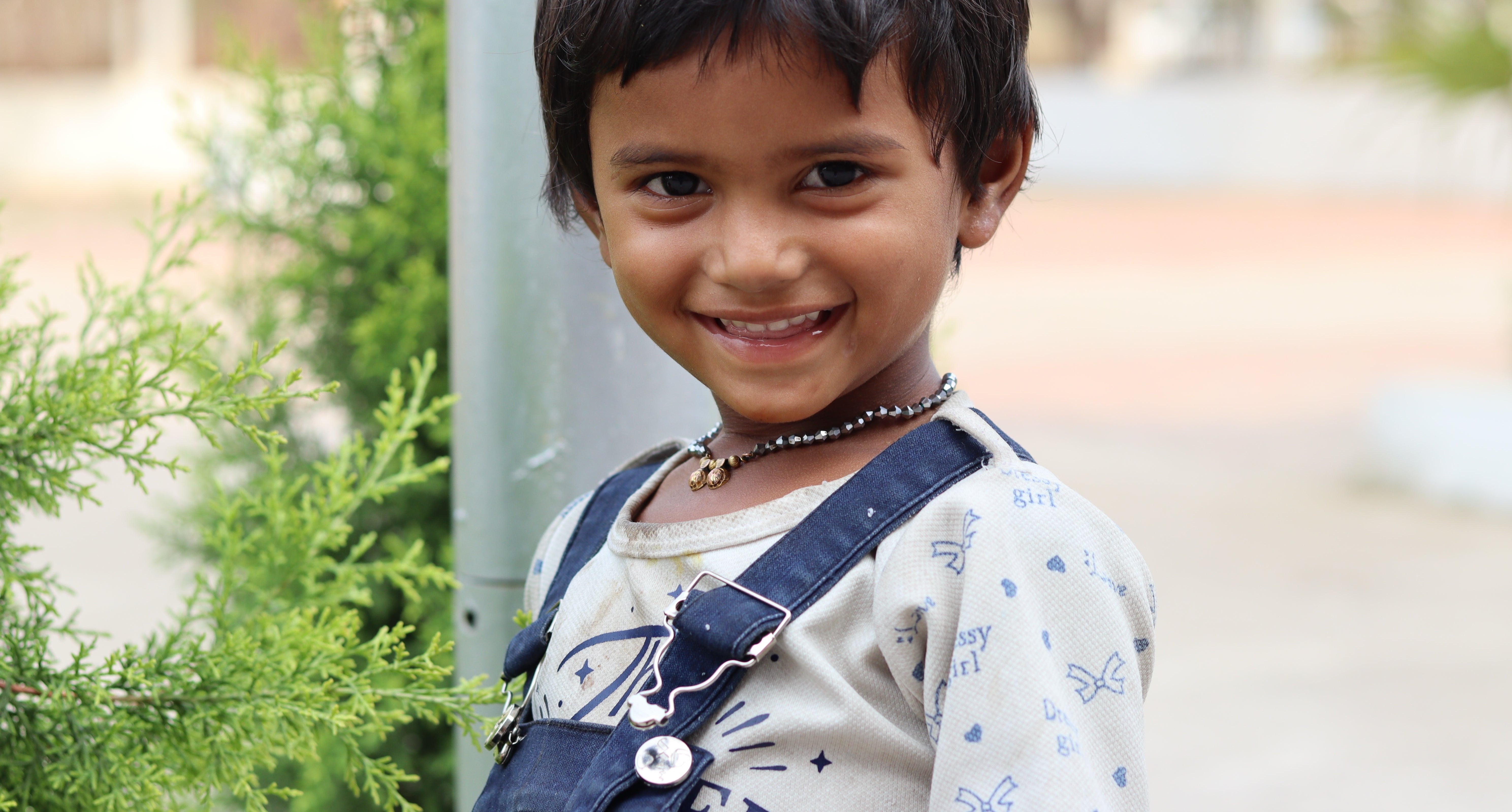 A young boy with black hair smiles warmly at the camera while standing outdoors next to green plants. The boy is wearing a light-colored shirt with printed patterns and denim overalls, and a beaded necklace. The background is softly blurred.