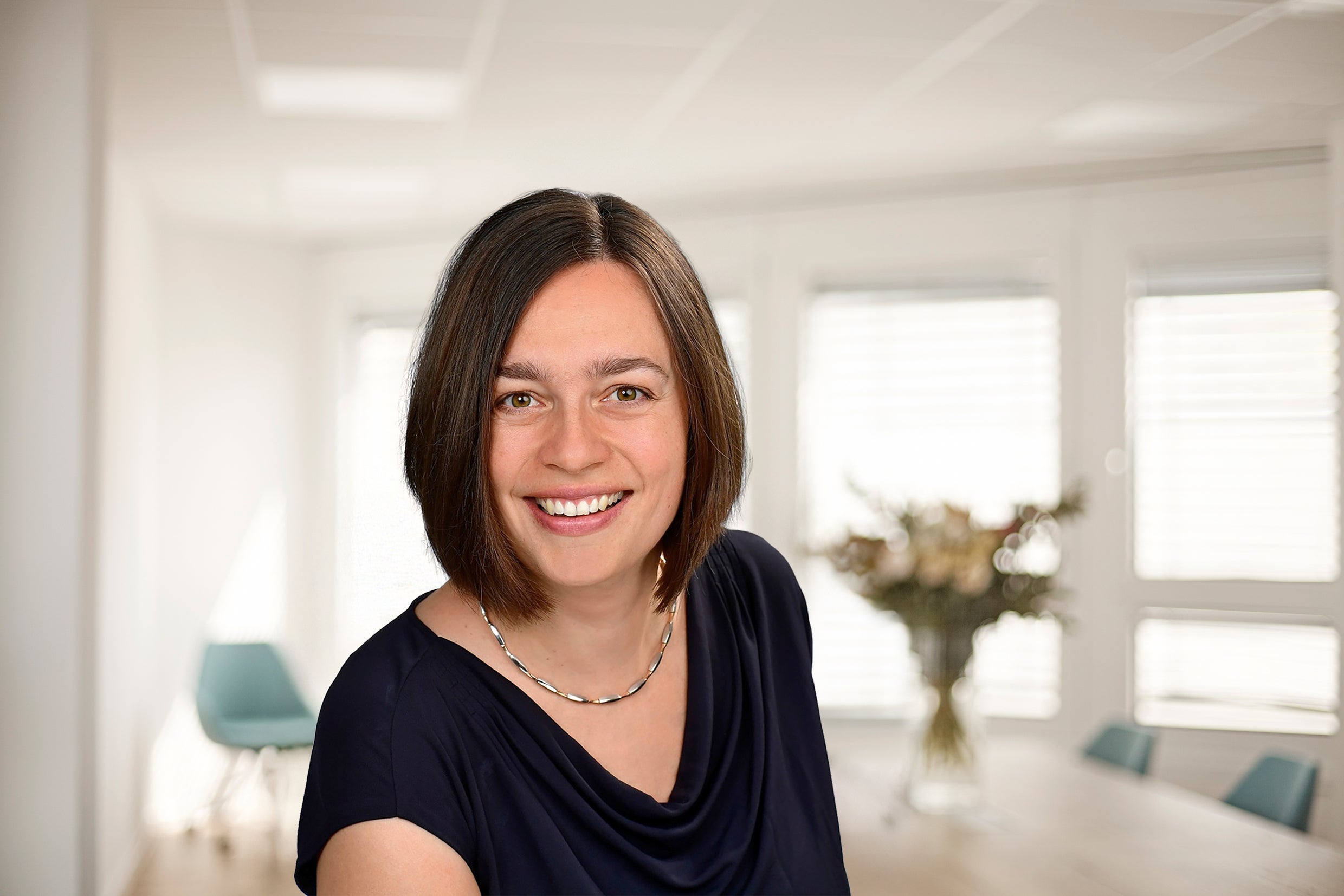 Portrait of Dr.Julia Pingel, Chief Information Officer, standing in a bright office space. She is wearing a black shirt, a and necklace, and is smiling at the camera. In the background is a bouquet of flowers on a office table, blue office chairs, and windows.
