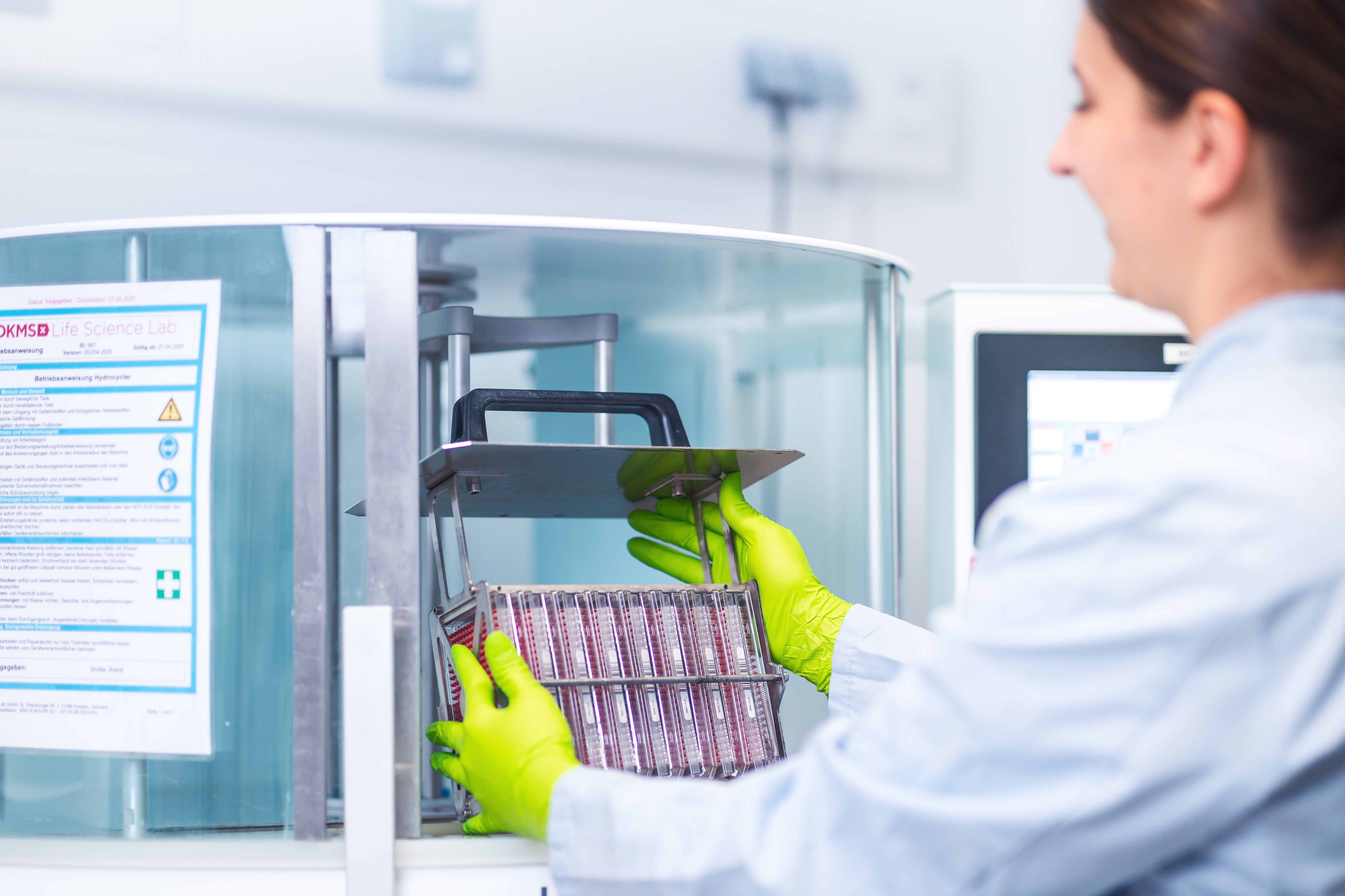 A lab technician wearing green protective gloves and a light blue lab coat is placing a metal rack filled with labeled sample containers into a laboratory device. A DKMS Life Science Lab instruction sheet is visible on the glass of the laboratory device, and a computer screen is in the background.
