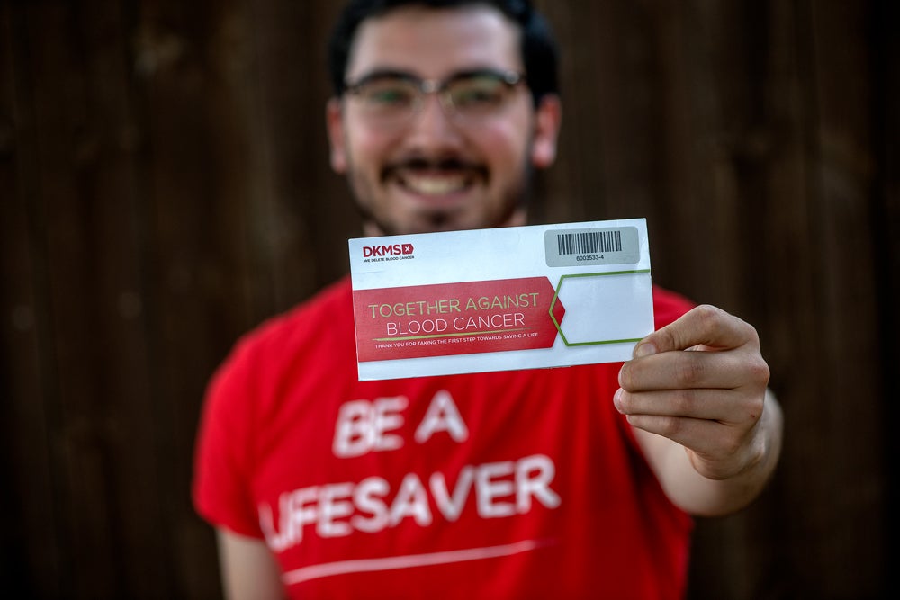 A smiling man with dark short hair and a beard, wearing glasses and a red "BE A LIFESAVER" T-shirt, holds a DKMS registration kit toward the camera with his left hand. His face and upper body are slightly blurred in the background, while the kit and his hand are in sharp focus in the foreground.