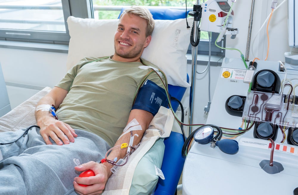 A young male donor, wearing a khaki-colored shirt and gray sweatpants, is lying comfortably on a medical chair, smiling at the camera while donating stem cells through peripheral blood stem cell (PBSC) donation. He is connected to an apheresis machine via tubes, and holding a red squeeze ball in his hand to support blood flow. In the background parts of windows can be seen.