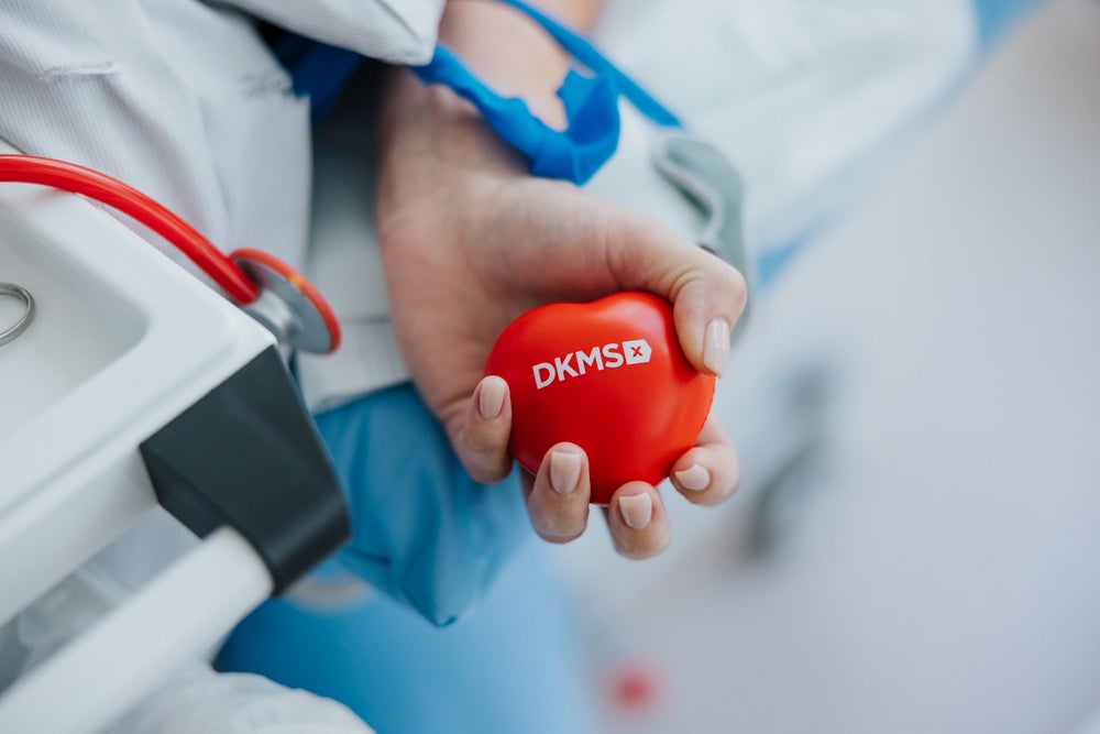 A close-up of a donor's hand holding a red heart-shaped stress ball with the DKMS logo during a stem cell donation. Medical tubes and equipment are visible in the background.