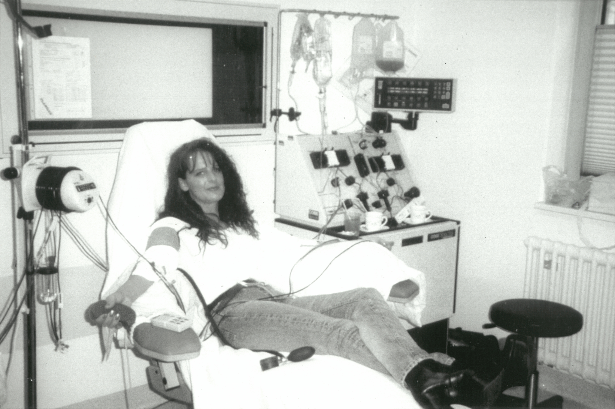 Black-and-white photo of a woman donating periphal blood stem cells while reclining in a medical chair. She is connected to an apheresis machine, holding a stress ball in one hand, and smiling slightly toward the camera.