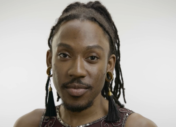 A portrait of the donor Deonté Lee. He has long, dark dreadlocks tied back in a braid, wears large hoop earrings, and a dark top. He's standing in front of a plain, light background, and looks confidently into the camera.