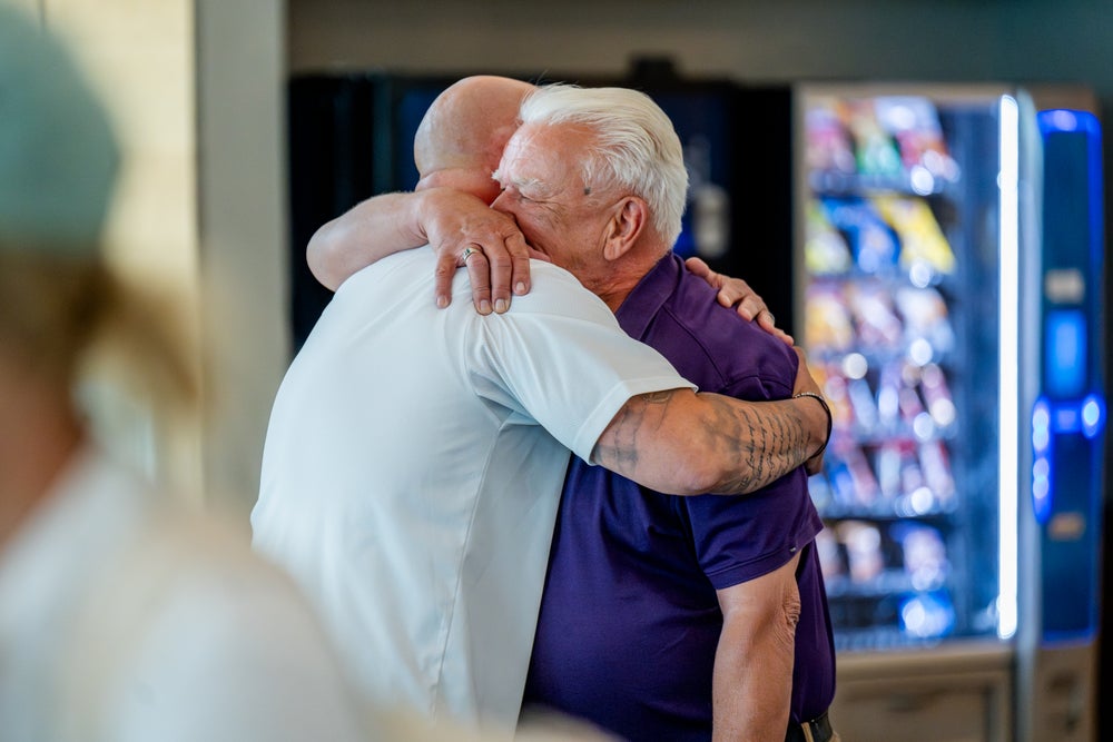 An emotional moment as two patients embrace tightly. One bald man, on the left, wearing a white shirt and has tattoos on his right arm; the other man, on the right, with white hair and a purple shirt, is closing his eyes during the hug. A vending machine is visible in the background.