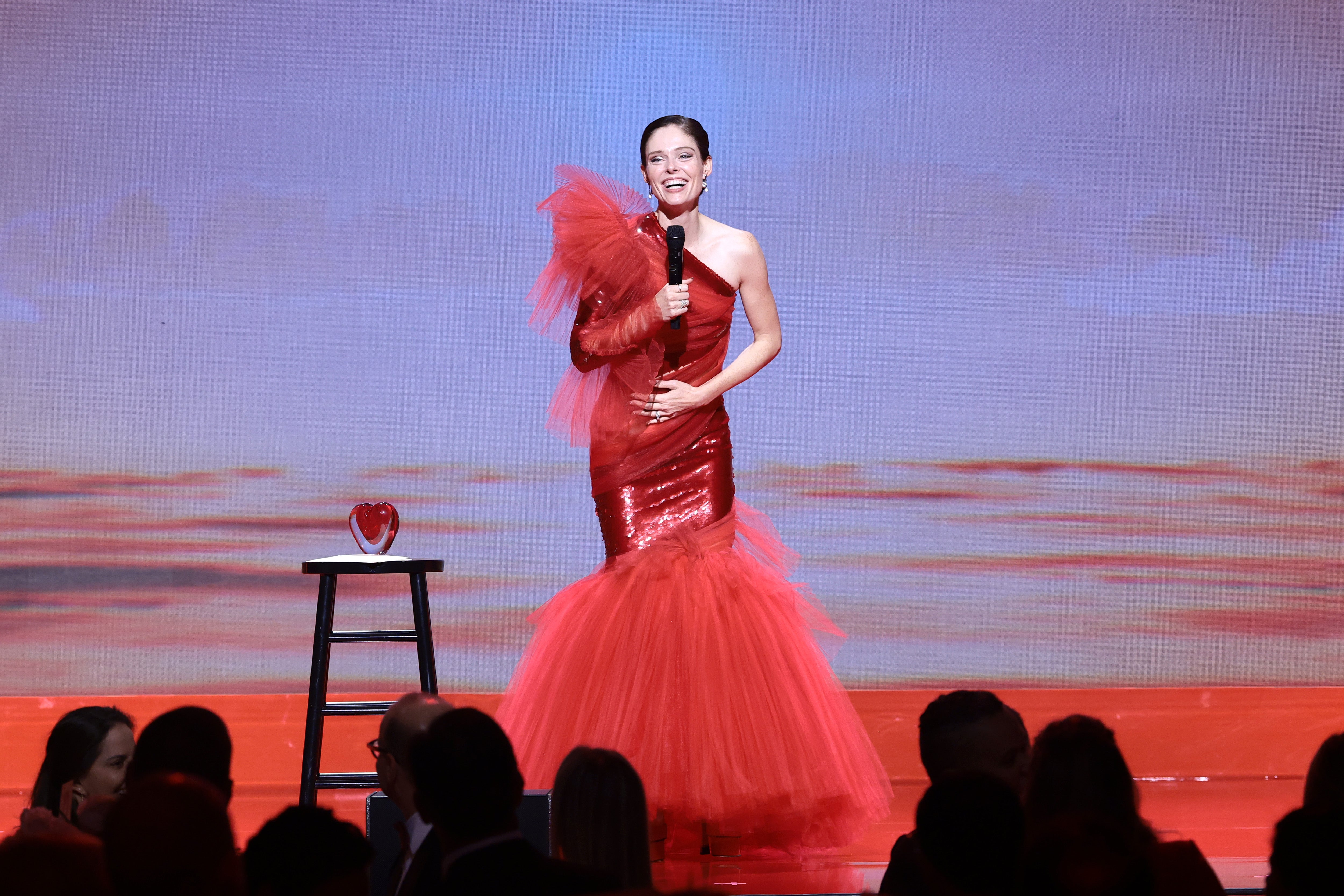 The DKMS Gala Honoree Coco Rocha in a red gown with tulle details, stands on stage at the DKMS New York Gala 2025 holding a microphone, and smiling as she addresses the audience. A red heart-shaped award sits on a stool beside her. The background shows a warm blue-red color gradient.