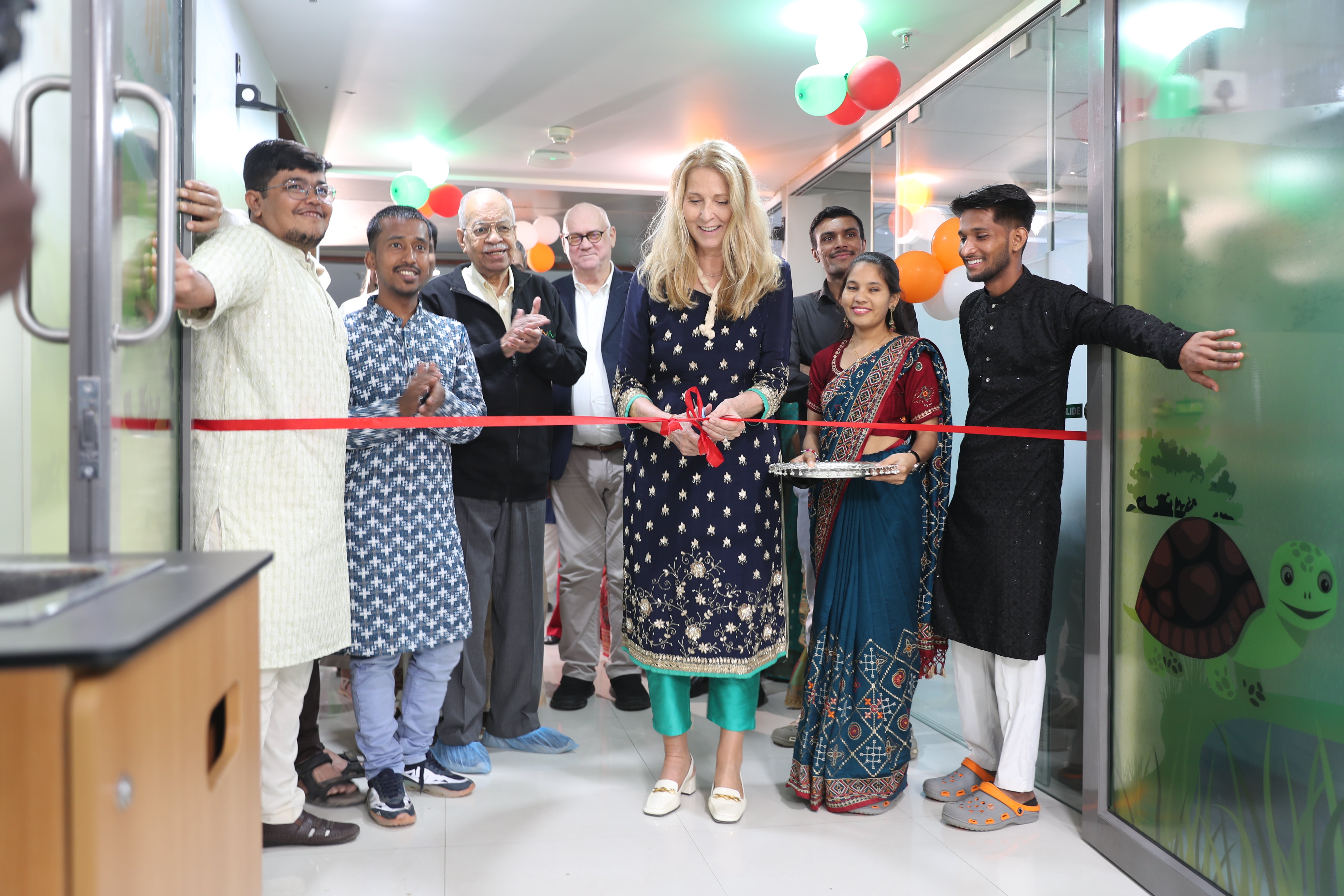 The CEO of DKMS Dr. Elke Neujahr, wearing a dark blue traditional outfit with golden applications, is standing in the middle of a group of people and cutting a red ribbon. They are celebrating the inauguration of the new bone marrow transplant unit for children in Ahmedabad, India. In the background the BMT unit hallway is decorated with balloons.