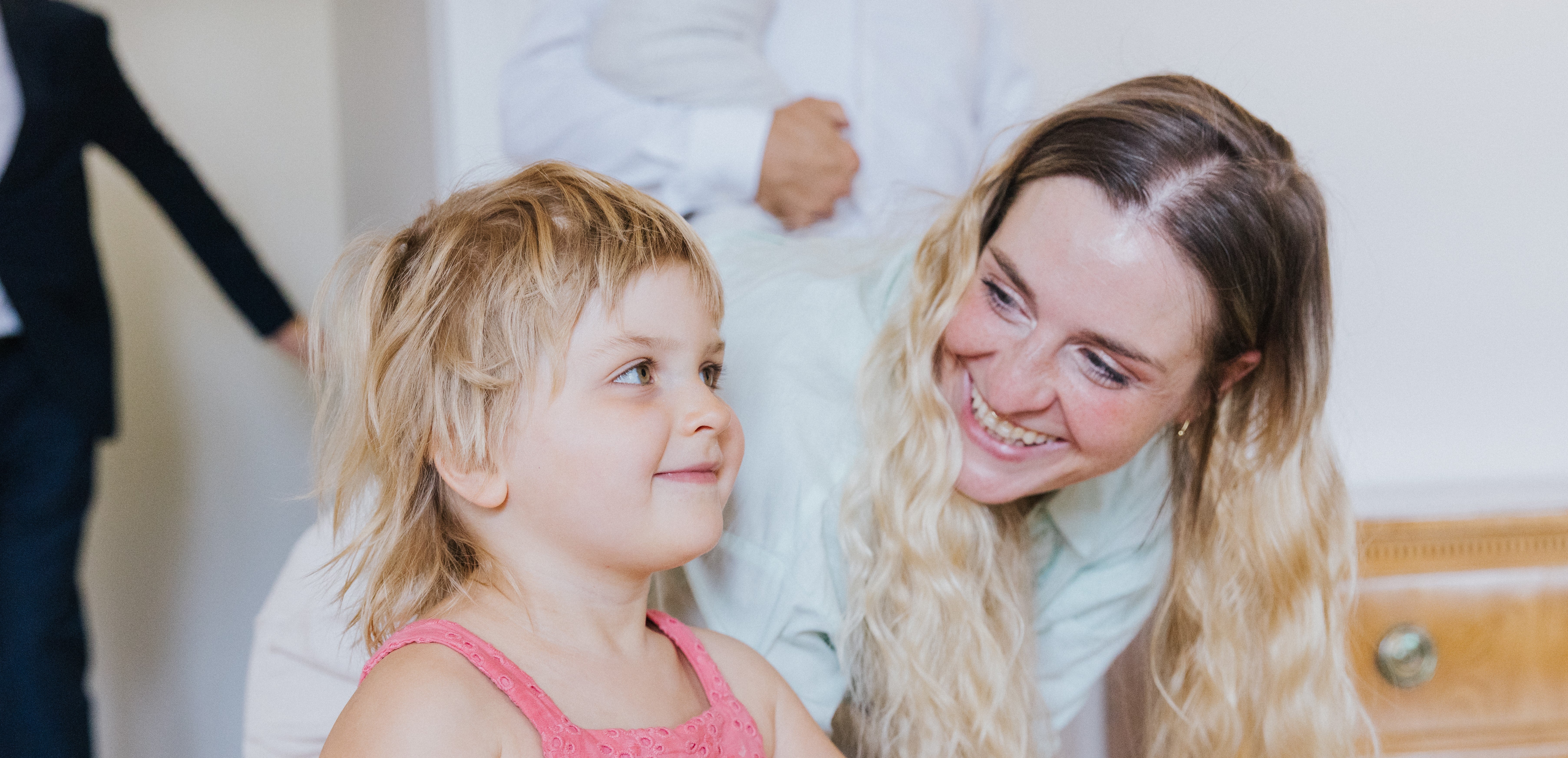 A smiling woman with long blond hair is bending down and looking at her little daughter with blond hair and a pink dress. The girl is smiling softly and looking slightly to the right side. The mood is warm and joyful, with other people blurred in the background.