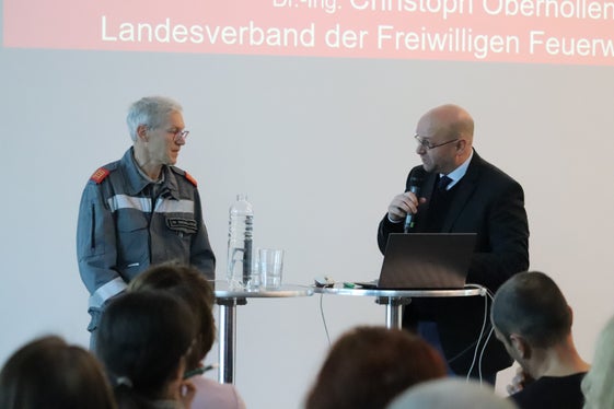 Il ruolo dei vigili del fuoco nella protezione dei Beni culturali: il moderatore della conferenza Gustav Pfeifer (a destra) passa il microfono a Christoph Oberhollenzer, direttore dell'Unione provinciale dei Corpi dei vigili del fuoco volontari dell'Alto Adige e direttore della Scuola dei vigili del fuoco (Foto: ASP/Maja Clara)