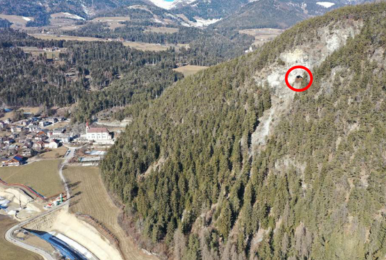 Il fianco del versante a rischio di caduta massi (il Koflloch è cerchiato in rosso) con il cantiere della circonvallazione di Chienes e la barriera provvisoria di protezione dalla caduta massi già eretta durante la fase di costruzione. (Immagine da drone: Ufficio tecnico strade Nord-Est)