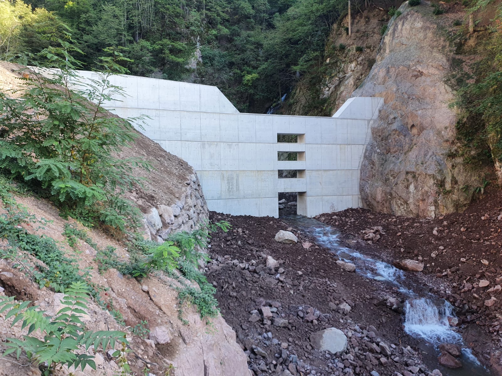 La nuova barriera di contenimento lungo il rio Sinigo è stata ultimata: la struttura è alta 12 metri nella sezione di uscita e 15 metri nell'ala laterale, è larga circa 40 metri e ha uno spessore in cemento armato di due metri e mezzo alla base (Ufficio sistemazione bacini montani Sud)