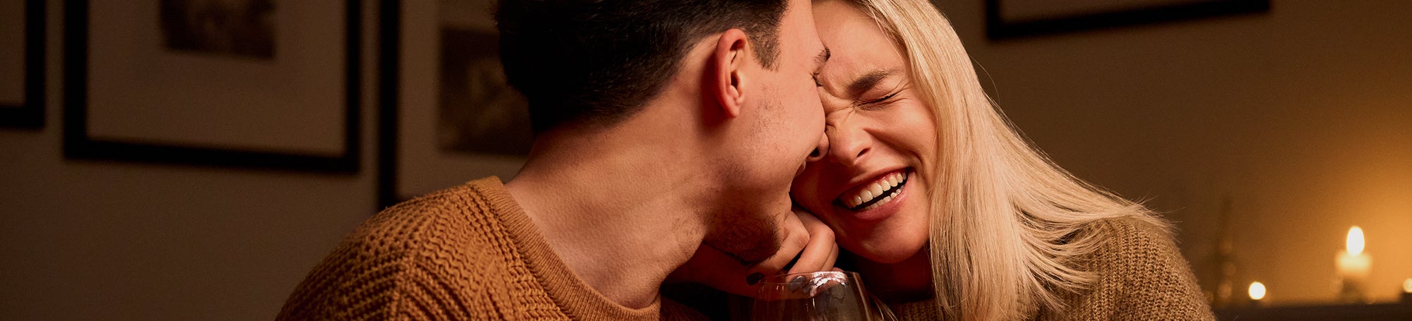Ein verliebt lachendes Pärchen sitzt bei Kerzenlicht am Esstisch einer Wohnung mit einem Glas Rotwein