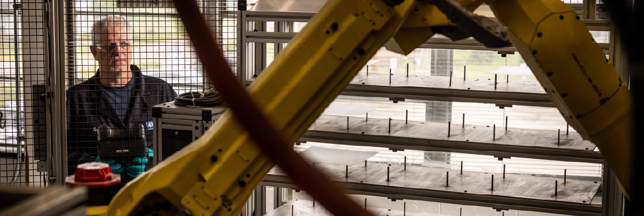 A technician observes a yellow industrial robotic arm operating inside a manufacturing enclosure.