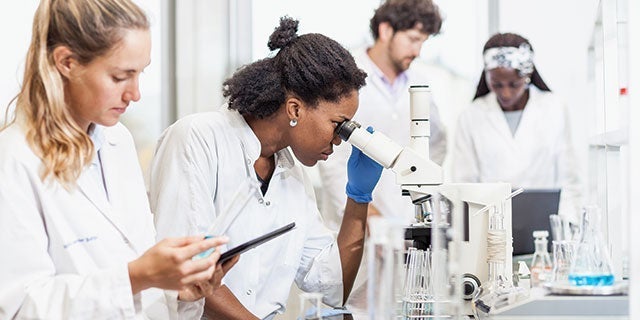 Group of scientists focusing on various tasks in a lab, one using a microscope, another analyzing data on a tablet