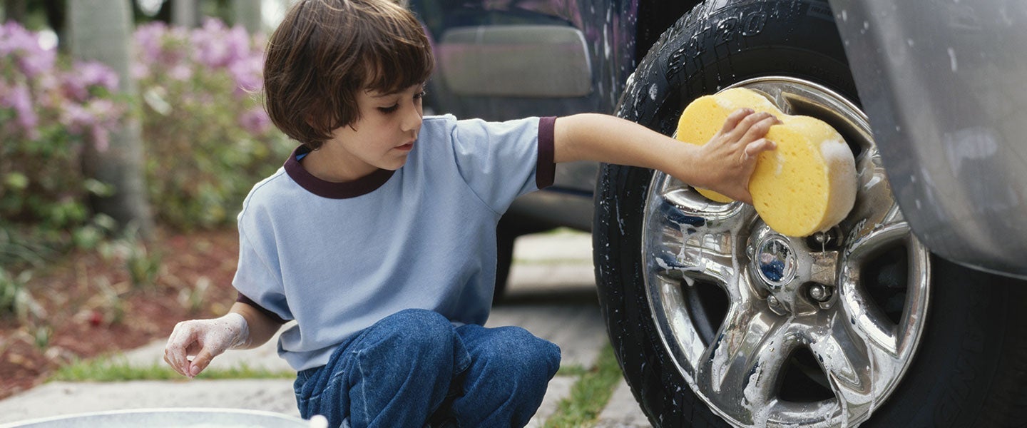 Young child in a blue shirt and jeans kneeling and cleaning a shiny car wheel with a yellow sponge, surrounded by greenery and pink flowers