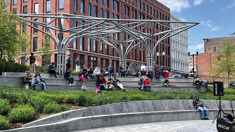 People relaxing and socializing in an urban park with modern steel structures and tiered seating areas, surrounded by historic brick buildings
