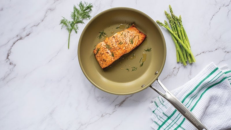 Cooked salmon fillet seasoned with herbs in a non-stick frying pan, accompanied by fresh asparagus and a sprig of dill on a marble countertop