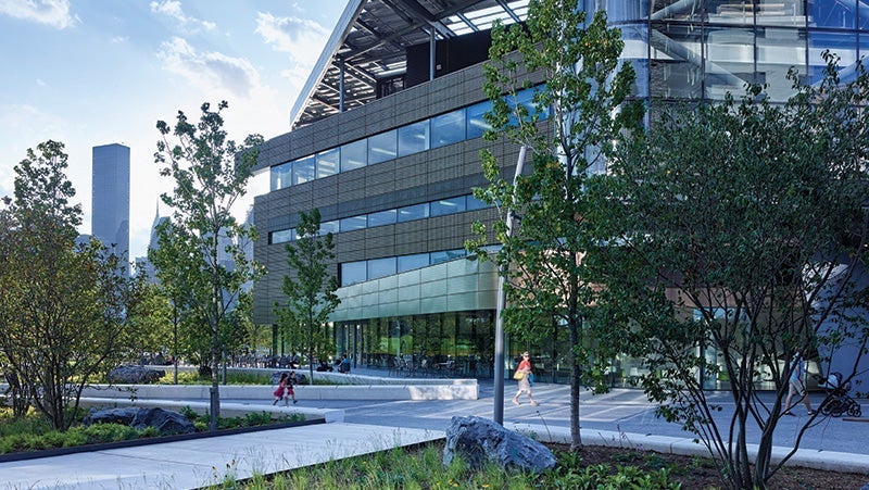 Modern glass and steel building with lush green landscaping, pedestrians, and a cyclist on a sunny day, featuring a backdrop of a city skyline