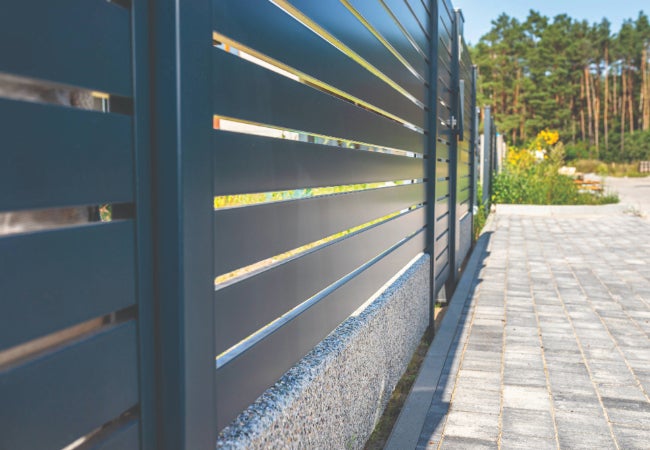Grey metal powder coated fence in an open paved area with green trees in the background