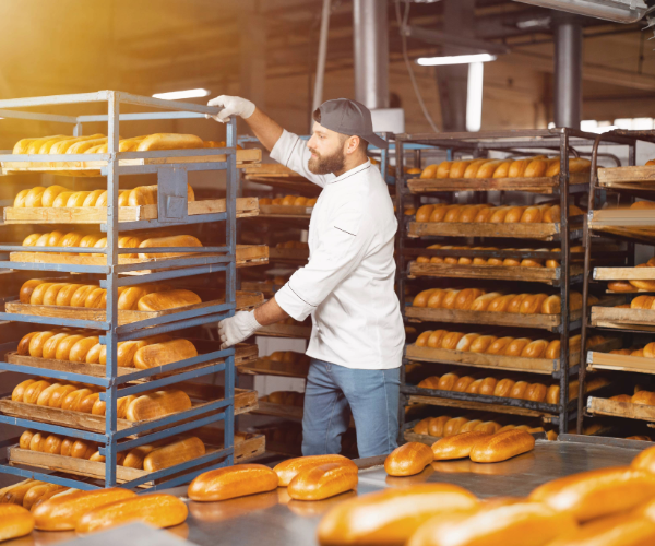 Bakery with multiple trays of baked baguettes and man in white jacket wearing white gloves