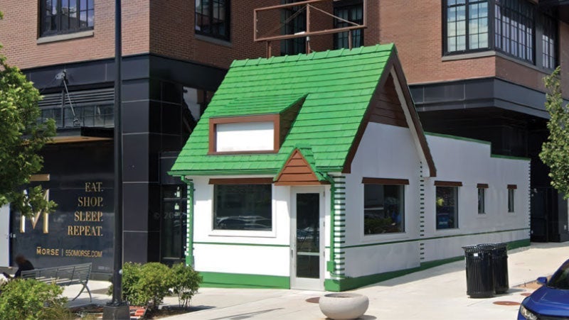 A small quaint house with bright green roof and white walls located on a city street, nestled between a modern building and a street lined with cars
