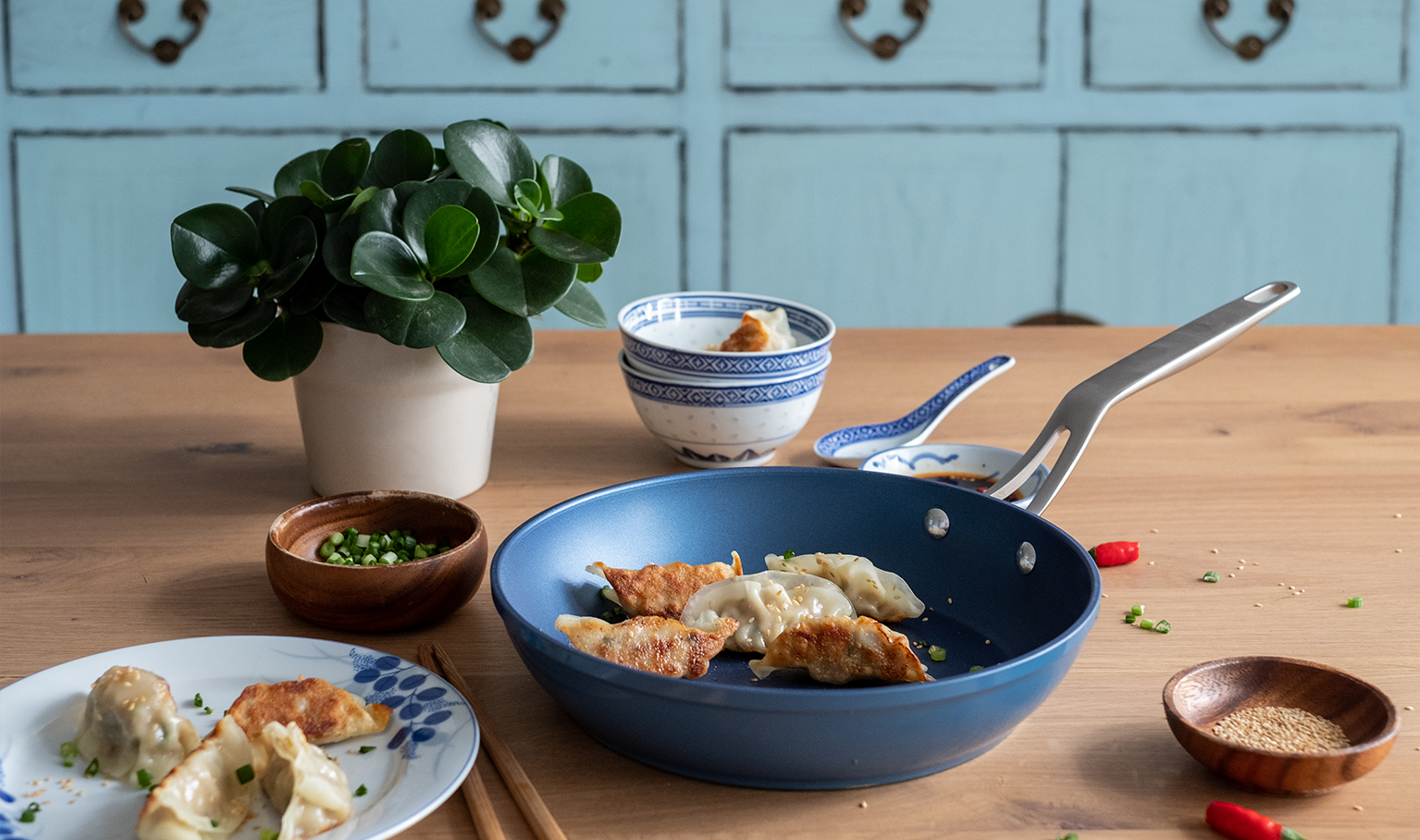 Blue Eterna coated pan on a wooden kitchen table holding oriental food with bowls in the background
