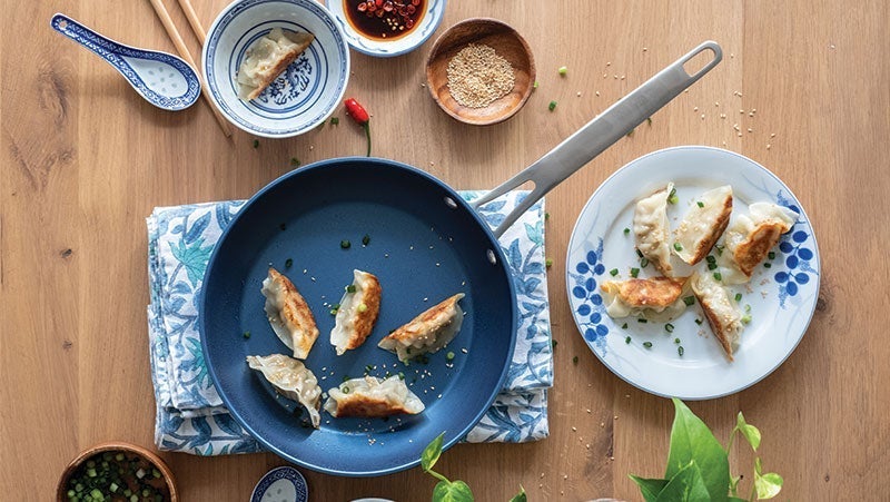A wooden table with pan-fried dumplings on a blue skillet and a white plate, accompanied by condiments and sauces in small dishes.
