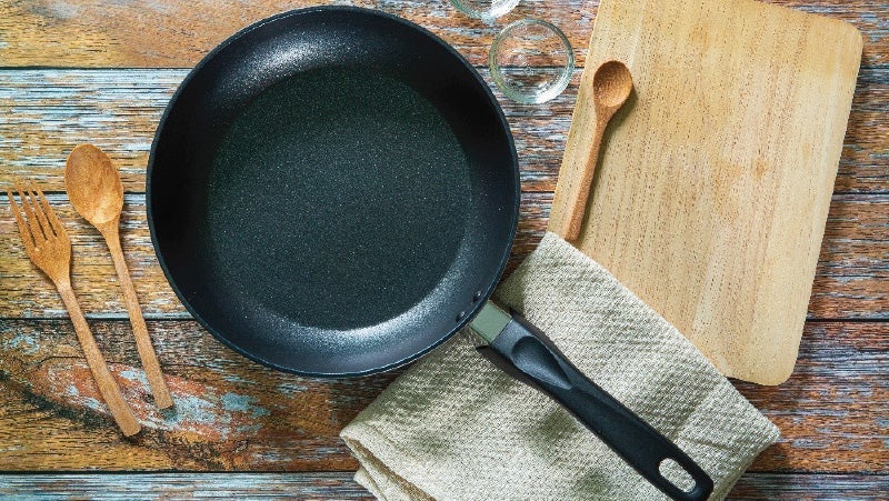 Non-stick skillet alongside wooden spoon, fork, and cutting board on a rustic wooden surface with a linen cloth