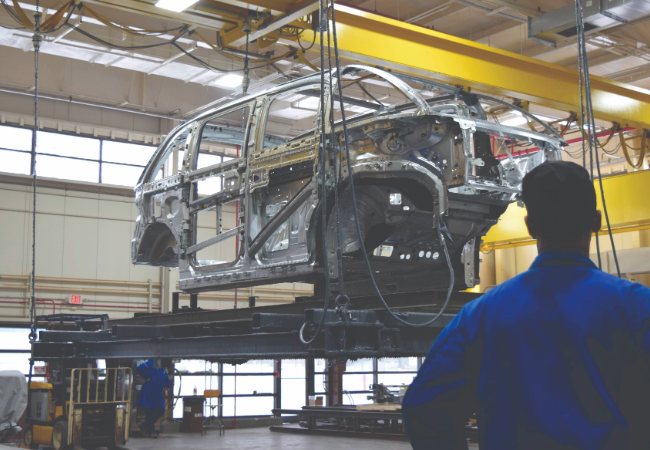 Metal car chassis on a yellow pretreatment line with a worker in blue overalls looking on 