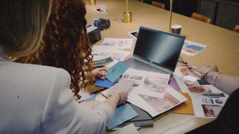 Over-the-shoulder view of a woman with curly hair at a conference table, reviewing various photos and documents with a colleague, with a laptop open in front of them