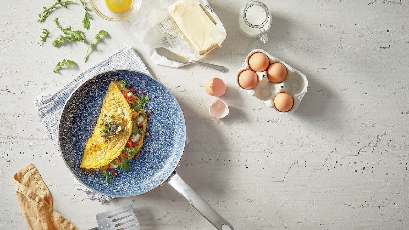 Top view of a vegetable-filled omelet in a blue speckled frying pan, surrounded by ingredients like eggs, butter, milk, and bread on a light textured surface