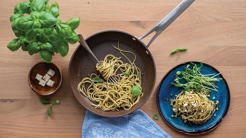 Overhead view of a wooden table featuring a skillet with pesto pasta, a blue plate with pasta and microgreens, beside a bowl of diced tofu and a basil plant
