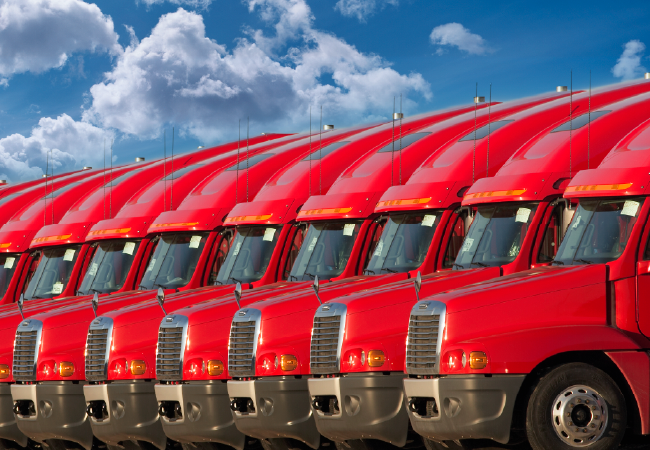 Row of red semi-trucks parked side by side, aligned uniformly under a partly cloudy blue sky.