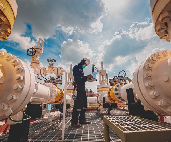 Worker wearing full PPE and checking documents alongside industrial pipework